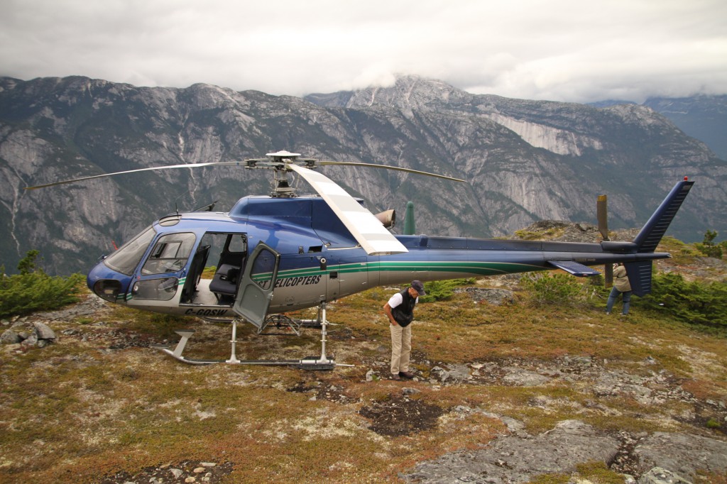 A man stands next to a helicopter perched on a mountain top.