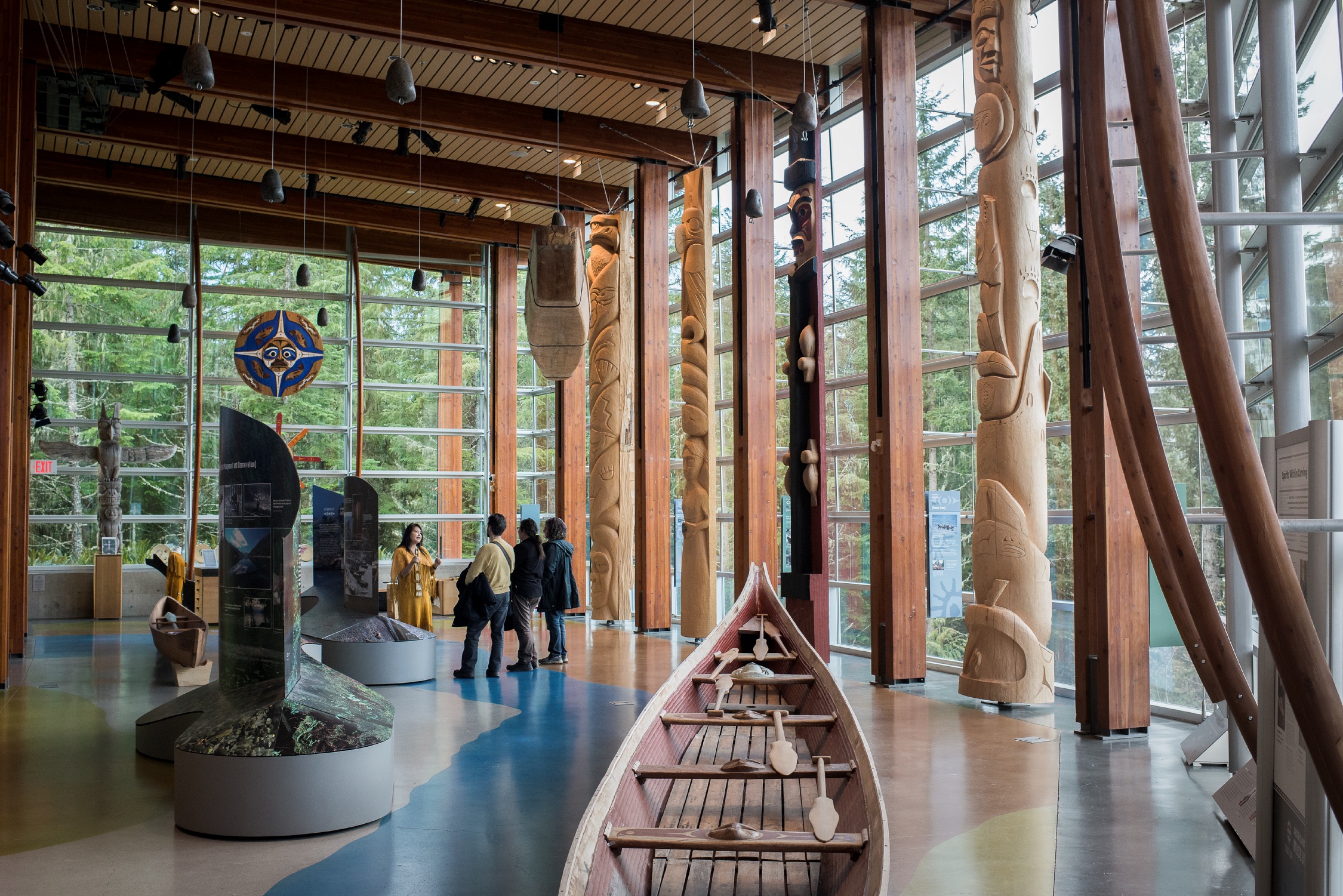 A group of visitors receive a tour of the SLCC. They are in a room with Indigenous works of art including totem poles and a canoe. 