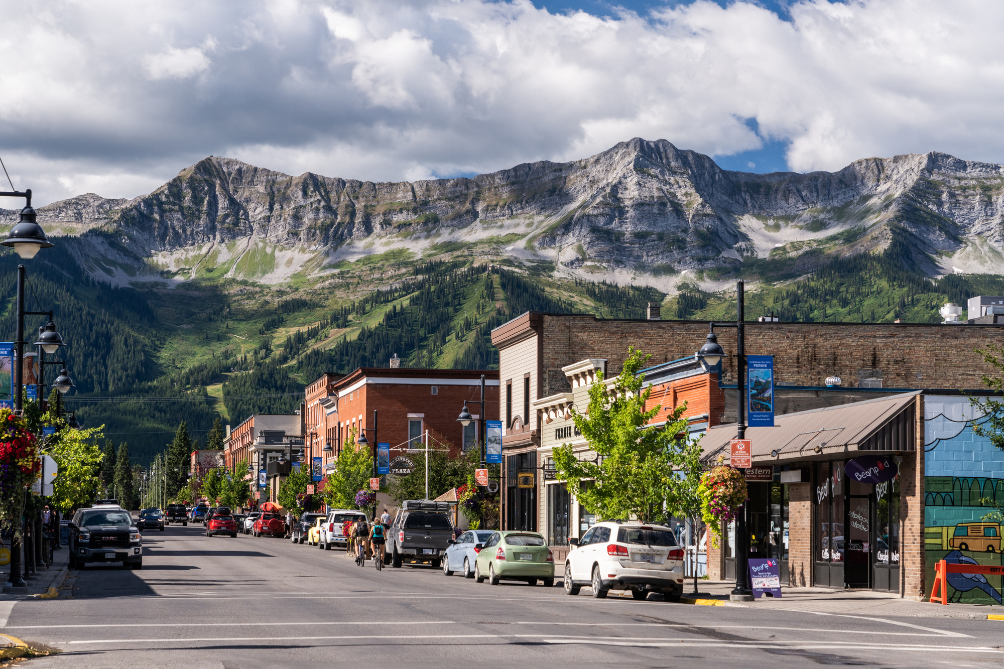 Cars are parked along a quiet street lined with heritage buildings. The sky is cloudy, and a dramatis mountain range is visible in the backdrop of the town.