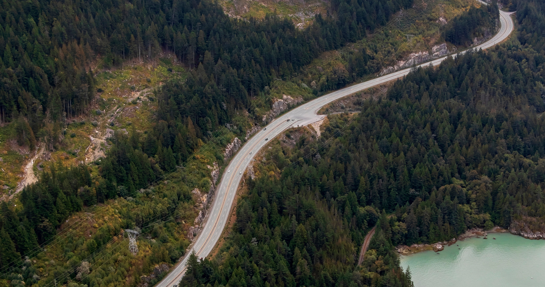 Two-lane stretch of Sea-to-Sky highway, hugging the tree-lined coast along Howe Sound, with islands visible in the background.