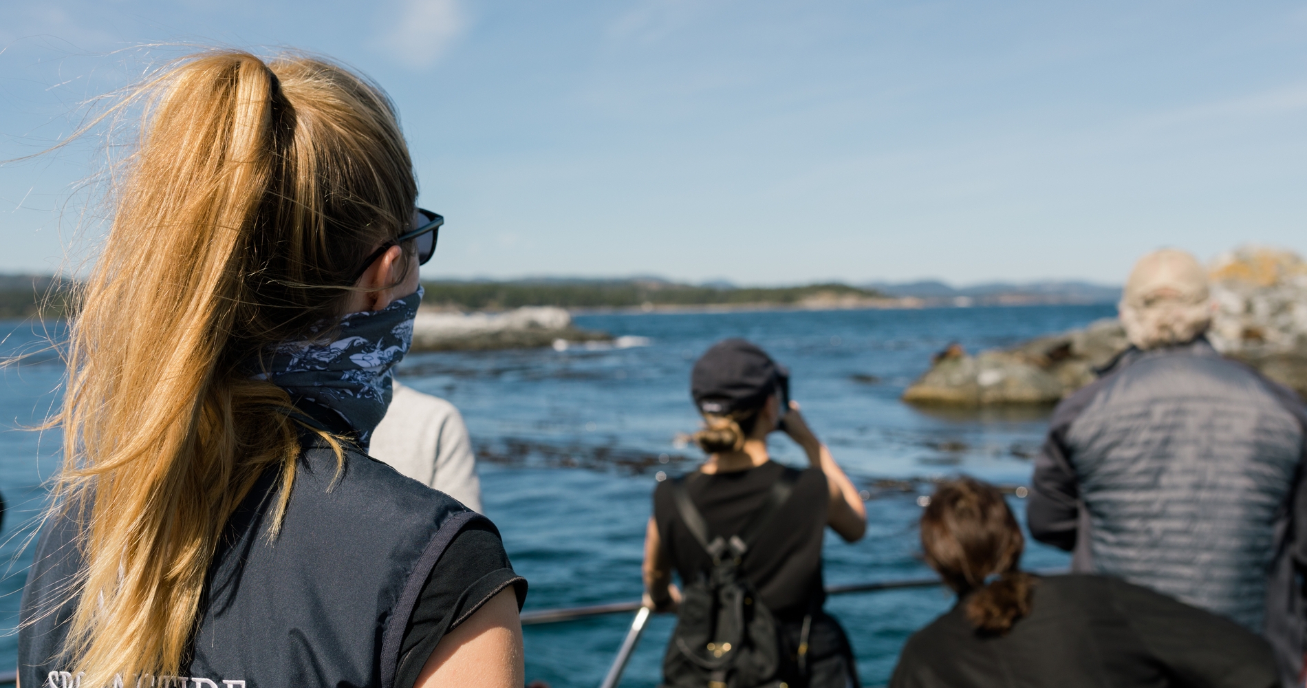 A group of people stands on a boat, looking out at the ocean and distant shoreline. The focus is on a person with long blonde hair in a ponytail, wearing sunglasses. Other individuals are taking photos or observing the scenery, enjoying the clear, sunny day. The water is calm and blue, with rocky outcrops visible in the distance.
